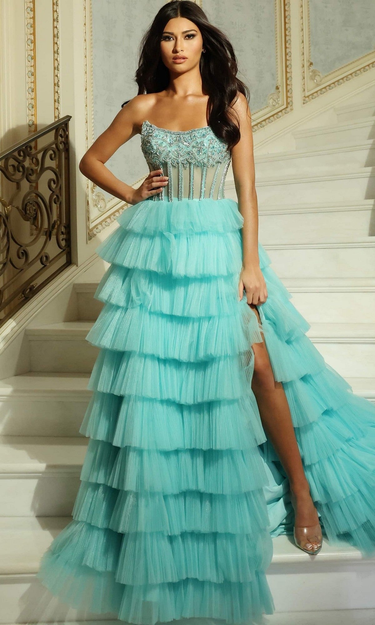 Woman stands on a staircase wearing the Jovani 44134 long aqua prom dress, featuring a strapless embellished bodice, ruffle-tiered tulle skirt, and high slit showing one leg.